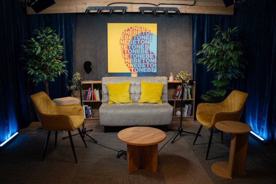 Studio 1 from the front in blue light: a grey sofa, two mustard armchairs, two microphones and a round wooden table.